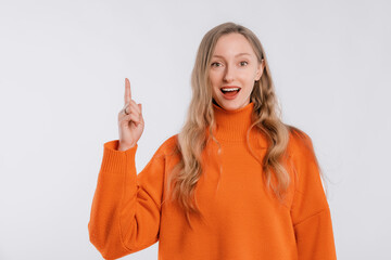 Smiling attractive young woman points finger up, shows way to banner, demonstrates logo or advertisement on top, stands in an soft orange sweater over white background