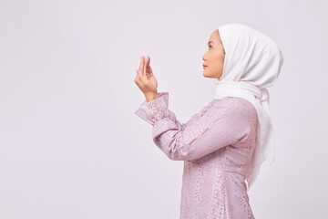 Portrait of young Asian Muslim woman wearing hijab and purple dress standing with raising hands and praying to god isolated over white background