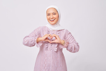 Portrait of smiling beautiful young Asian Muslim woman wearing hijab and purple dress showing heart...