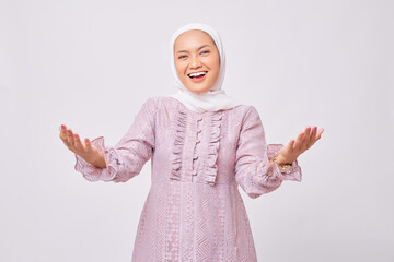 Portrait of excited beautiful young Asian Muslim woman wearing hijab and purple dress looking at camera and spreading her hands, welcoming Ramadan isolated on white studio background