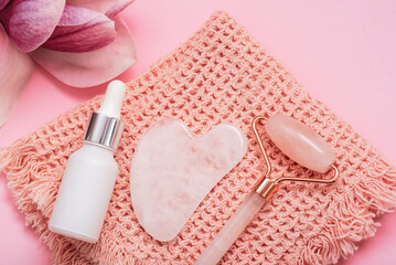 Gua sha stone, face roller, cosmetic products and magnolia flower on towel on pink table. Top view,...