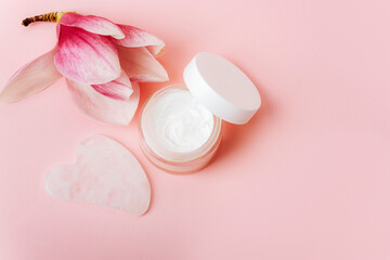 Obraz na płótnie Canvas Cosmetic cream jar and Gua sha stone for beauty facial massage therapy. Flat lay on pink background with magnolia flowers. Copy space