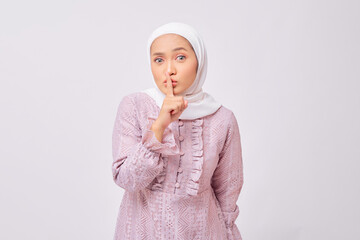 Portrait of serious beautiful young Asian Muslim woman wearing hijab and purple dress showing silence gesture, holding finger near her lips and looking camera isolated on white studio background