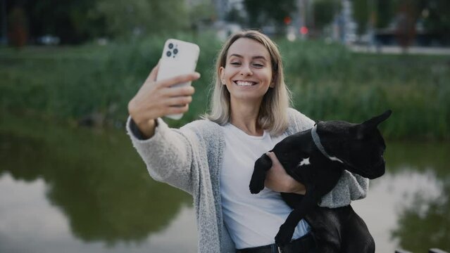 Beautiful Happy Woman is Standing Near the Local Lake with her Lovely Black French Bulldog and Doing Selfie, Making Memories with Her Puppy. Technology and People Dogs Friendship Concept