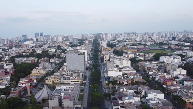Drone flying forward above a street called "Avenida Benavides" with many trees in the middle of the 2 lanes, going into the distant horizon of the city. Located in Miraflores district of Lima, Peru.