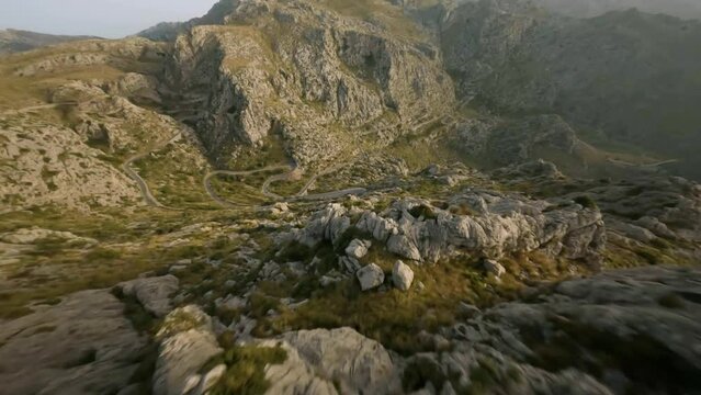 FPV drone shot flying downhill over winding Sa Calobra road along rocky mountain range in Mallorca, Spain during evening time.