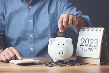 Financial calculate and planning, Clever yearly plan for money saving, Man holding coin and add to white piggy bank with coin pile and 2023 desk calendar on the table. Studio shot on dark backdrop.