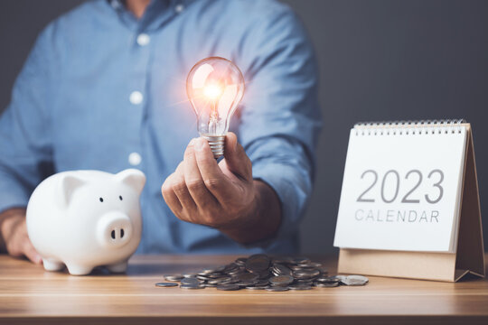 Financial calculate and planning, Clever yearly plan for money saving, Man holding light bulb with piggy bank, coin pile and 2023 desk calendar on the table. Studio shot on dark backdrop