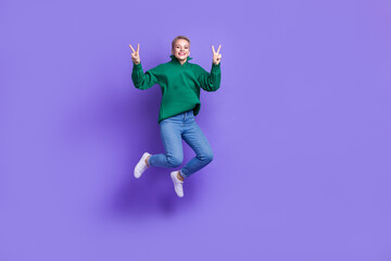 Fototapeta na wymiar Full length photo of pretty excited lady wear green sweater jumping showing v-sign empty space isolated violet color background