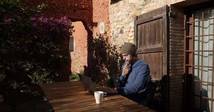 A man sitting at the laptop outside a country house, touching his goatee whilw thinking.