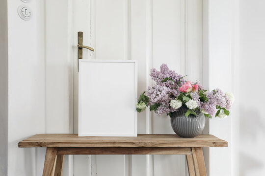 Vintage spring floral still life. Blank picture frame mockup on old wooden bench, table. Vase with bouquet of lilac, viburnum and tulips. White wall background. Empty copy space. Rustic interior.