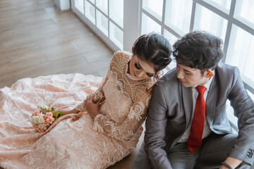 asian bride leaning on the groom while sitting together on the floor beside the window as the...