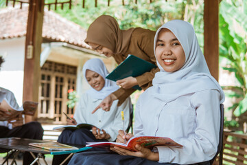 Middle school girls wearing headscarves smiling holding pens and books sitting in the gazebo during outdoor class