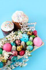 Easter cakes, colorful eggs, cherry flowers in carton box on abstract blue background. Happy Easter russian text greeting. Easter holiday concept. spring festive season.