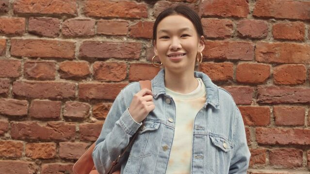 Slowmo tilt up portrait of Asian female first year college student in oversized denim jacket posing for camera standing against red brick wall outdoors