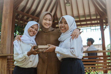 female teacher in hijab and two female students smiling while giving gifts outdoors