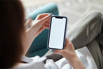 Mock up white screen blank mobile phone in woman hands holding sitting on armchair at home Back view