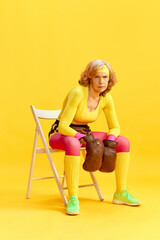 Middle-aged sportive woman in colorful uniform sitting on chair in retro boxing gloves with serious face against yellow background. Concept of sportive lifestyle, retirement, health care, wellness. Ad
