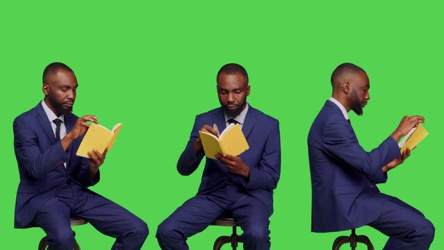 African american person reading pages of story book, sitting over greenscreen background. Office employee feeling happy reading literature novel in business suit, confident man.