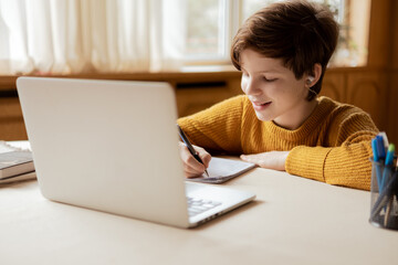 A smiling child writes down an assignment in a notebook from a teacher at an online school. Comfortable learning at home