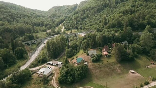 Dynamic aerial view with FPV racing drone flying above cottages and under an old railway bridge alongside an empty winding road. LuPa Creative.