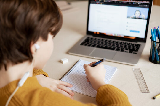 The Boy Sits In Class On His Laptop At An Online School And Watches As The Teacher Explains The Material And Writes It Down In His Notebook. Back View Of The Screen