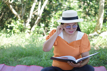 Asian middle- aged woman sits at the park, reads book, holds eyeglasses, has difficulties to see...