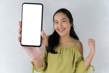 Asian girl in a cheerful green shirt points to a handful of invisible objects. pose over a gray background. Showing mobile phone with empty screen, mockup.