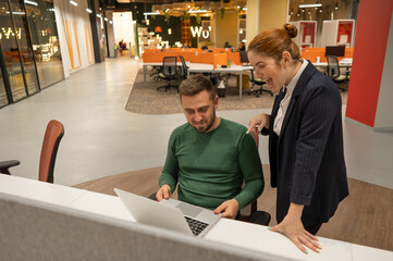 A red-haired Caucasian woman stands at the desktop of a bearded man. The boss approves the work of the subordinate. 