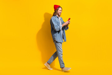 Naklejka premium Full size photo of positive man wear denim jacket jeans trousers hold smartphone walk empty space isolated on yellow color background