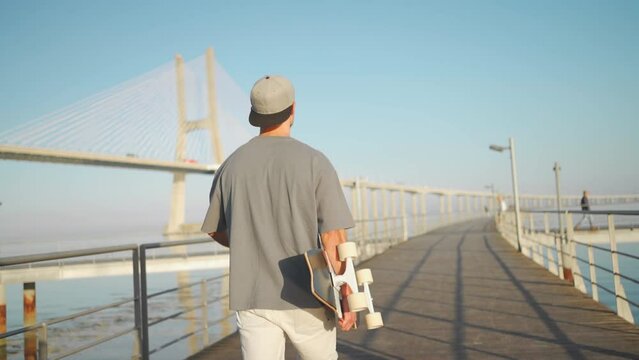 Man walk with skateboard on bridge summer day near ocean. Adult male person skateboarder enjoy after riding on skate. Millenial hipster with longboard 