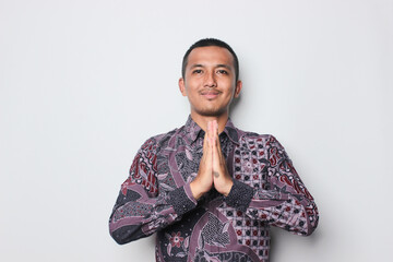 Smiling young Asian man wearing shirt gesturing traditional greeting isolated on white background.