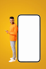 Smiling attractive indian guy in casual wear standing near huge smartphone with empty blank screen, looking at the screen and using phone, hipster man advertising new app or website, mockup