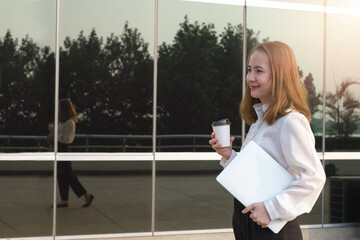 Professional business women going to work in the morning walking in the city building .She was smiling and her hand holding a laptop and hot coffee cup.