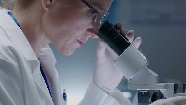 Experienced female scientist in gloves and lab coat looking in microscope when carrying out research in clinical laboratory. Side view close-up shot