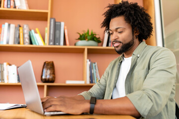 Friendly and positive african-american male employee or student using laptop, businessman looking at the screen, typing, responding to emails sitting in contemporary office
