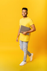 Cheerful Indian man carrying laptop computer isolated on yellow. Happy arab male student with laptop walking, freelancer using portative gadget, computer. Vertical photo