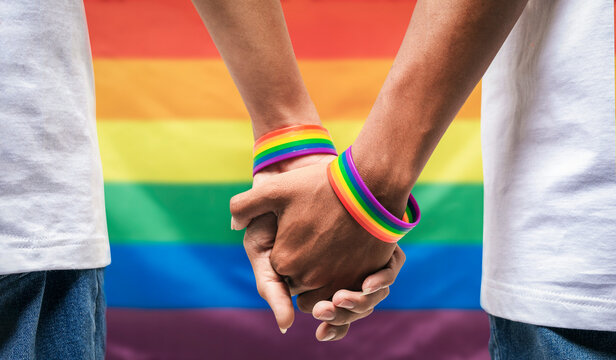 Male couple wearing gay pride rainbow awareness wristbands holding hands on rainbow flag background. lgbt, same-sex relationships and homosexual concept