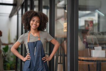 Portrait of African American businesswoman standing at the cafeteria door entrance. A cheerful young waitress in a blue apron near a glass door with an open signboard and looking at the camera.