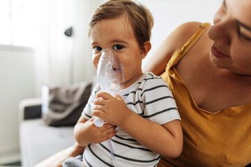 Portrait of a baby boy inhaling medicine in nebulizer with his mother at home. - obrazy, fototapety, plakaty