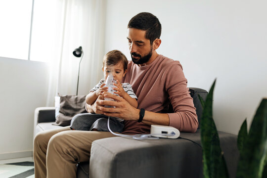 Portrait of a baby boy inhaling medicine in nebulizer with his father at home.