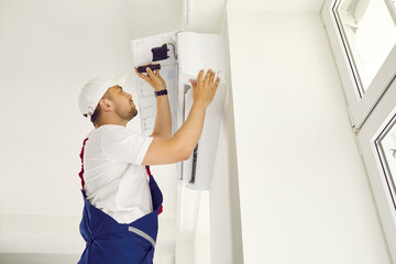 Young male worker uses a screwdriver while installing a new modern air conditioner unit on a white wall inside a house or an office building. AC installation, maintenance and repair service concept