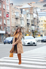 Fototapeta premium A modern brunette girl in a brown coat and high boots and a beige handbag walking across the road. Walk. Autumn. urban concept
