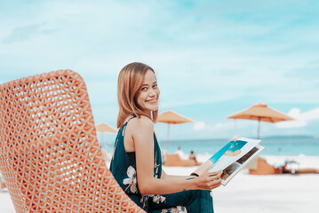 A young lady in a blue floral dress reading a lifestyle magazine while sitting on a comfy lounge chair near the beach. During her vacation at a tropical resort.