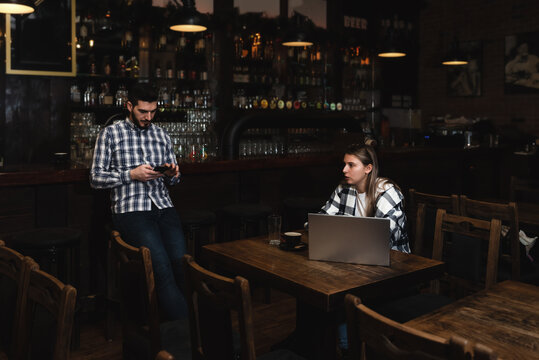 Two Diverse Entrepreneurs Have a Team Meeting in Their Stylish Coffee Shop. Barista and Cafe Owner Discuss Work Schedule and Menu on Mobile or Computer. Young Female and Male Restaurant Employees