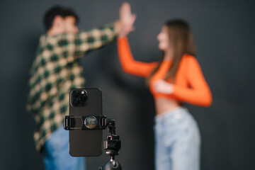 Selective focus on smartphone set on tripod over cheerful young couple high five isolated over black background. Creating and sharing content for social