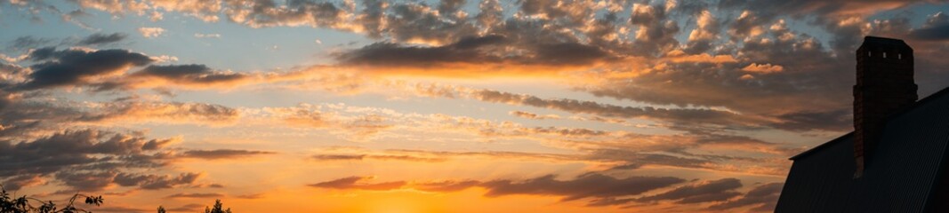 A summer sunset in yellow and orange colours. The sun's rays from below illuminate the dark clouds. In the foreground is a chimney on the roof of a house. Panoramic photo.