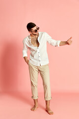 Full-length portrait of handsome young man in stylish clothes, white shirt and beige jeans posing against pink studio background. Cheerful mood. Concept of emotions, male fashion, lifestyle. Ad