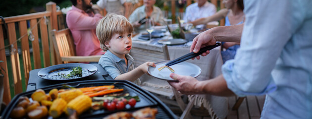 Father putting grilled meat and vegetable on plate to his son during family summer garden party.