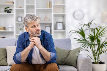 Adult mature man pensive sitting alone at home on sofa close up, gray haired person thinks looks...